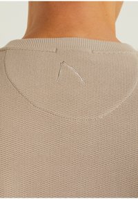 Detailní záběr na béžový pletený svetr, který ukazuje texturovanou látku, žebrovaný límec a malý vyšitý detail poblíž výstřihu.