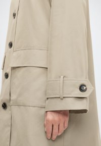 Μανίκι μπεζ καμπαρντίνας με μανσέτα με κουμπί και μπροστινή τσέπη, που δείχνει το χέρι ενός ατόμου χαλαρό στο πλευρό του.