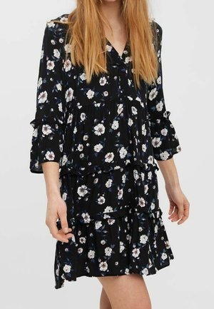 Kobieta w czarnej sukience w kwiaty z długimi rękawami i falbankami, stojąca na jasnym jednolitym tle.