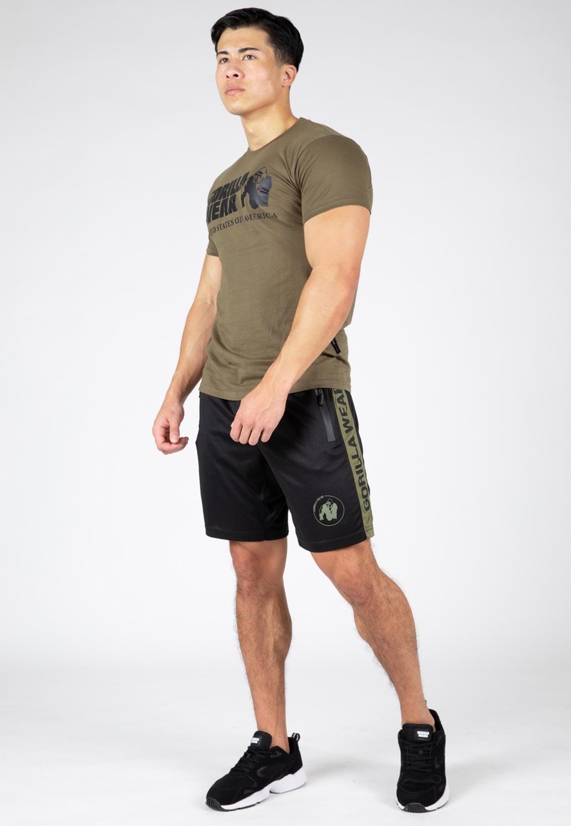 T-shirt verde a maniche corte con logo grafico nero e testo, abbinato a pantaloni corti neri con dettagli laterali verdi e tasca con zip. Scarpe sportive nere.