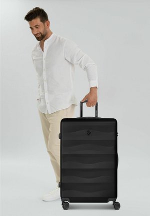 Uomo con camicia bianca e pantaloni beige che sorride mentre tiene la maniglia di una grande valigia nera con ruote.