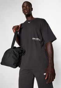 Juodas per didelis t-shirt su baltu tekstu „PUMA CREW“, antros spalvos kelnių; modelis laiko juodą sportinę krepšį su peties dirželiu.