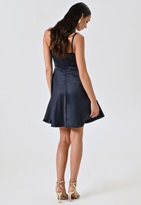 Robe en satin bleu marine avec un corsage ajusté, une jupe évasée et des bretelles spaghetti ajustables. Portée avec des talons dorés à lanières. Vue de dos affichée.