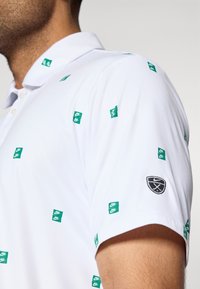 Λευκό polo μπλούζα με πράσινα λογότυπα της Nike σε διάσπαρτο μοτίβο, διαθέτοντας ένα μαύρο κεντημένο σήμα στο μανίκι και κλασικό σχέδιο γιακά.