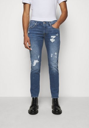 Man draagt slim fit blauwe versleten spijkerbroek, wit t-shirt en zwarte veterschoenen, staand met één hand in de zak.