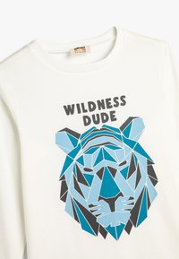 Weißes Langarm-T-Shirt mit einem geometrischen blauen Tiger-Design und schwarzem Text "WILDNESS DUDE" über dem Motiv. Weiches Material.