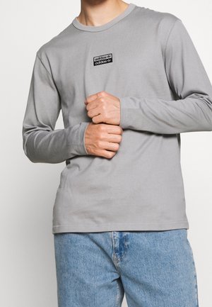 Mężczyzna w jasno-szarej, długorękawowej koszulce Adidas i niebieskich dżinsach, ręce splecione na talii na białym tle.