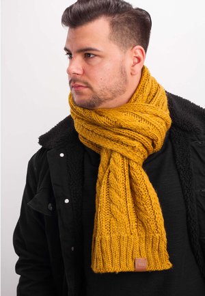 Gelber gestrickter Schal mit Kabelmuster, dicker Textur und gerippten Enden; verfügt über ein kleines Lederlabel; getragen über einer schwarzen Jacke.