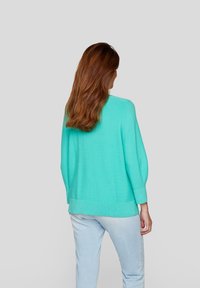 Türkiser Strickpullover mit langen Ärmeln, lockerer Passform, gerippten Bündchen und Saum, getragen über hellblauen Hosen, von hinten gesehen.