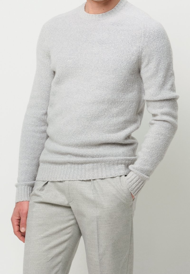 Homme portant un pull en maille gris clair et un pantalon gris clair assorti, une main dans la poche sur un fond uni.