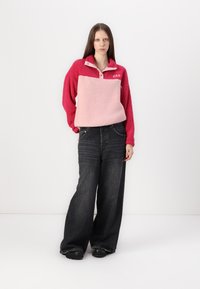 Fleece pullover in rood en roze met een kraag en een zak aan de voorkant, gecombineerd met een wijde spijkerbroek van donker denim en zwarte schoenen.