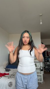 White ribbed tank top paired with light blue and white striped shorts. Long dark hair with teal streaks. Hands raised in a greeting gesture.