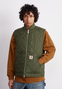 Quiltana olivno zelena brezrokavnika z visokim ovratnikom, sprednjimi žepi, zadrgo in logotipom Carhartt. Nosena čez rjav dolgrokavnik.