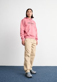 Roze hoodie met een voorzak en zwart logo, gecombineerd met losse beige broek en grijze sportschoenen. Staande op een blauwe vloer.