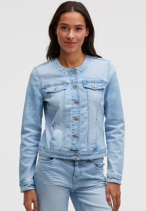 Frau trägt eine hellblaue Jeansjacke mit Knopfverschluss und passende Jeans, steht vor einem einfarbigen hellen Hintergrund.