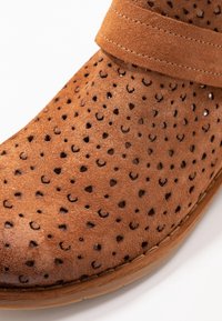 Bottine en daim marron avec un design perforé et une large sangle sur le dessus. Bout arrondi avec une semelle plate et des détails cousus.