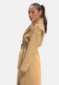 Cappotto lungo color cammello con cintura, ampi revers, silhouette slanciata, texture morbida e tasche laterali con spacco, mostrato di profilo laterale.