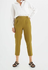 Olijf groene broek met een elastische taille, toelopende benen en zijzakken, gecombineerd met een wit shirt met kraag en zwarte sandalen.