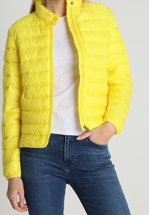 Kvinde iført en skarpt gul vatteret jakke over en hvid skjorte og blå jeans, med den ene hånd i lommen og jakken delvis lynet.