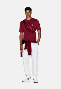 Bordeaux t-shirt, witte joggingbroek en sneakers met rode accenten. Zwarte schoudertas, maroon trui in laagjes om de taille geknoopt.