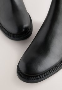 Čierne kožené členkové čižmy s hladkou textúrou, zaoblenou špičkou a jemným prešitým lemom pozdĺž podrážky, s nízkym podpätkom.