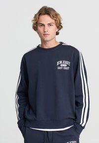 Felpa della marina in misto cotone con strisce bianche sulle maniche. Presenta una grafica "New Jersey East Coast" sul petto. Collo e polsini a coste.