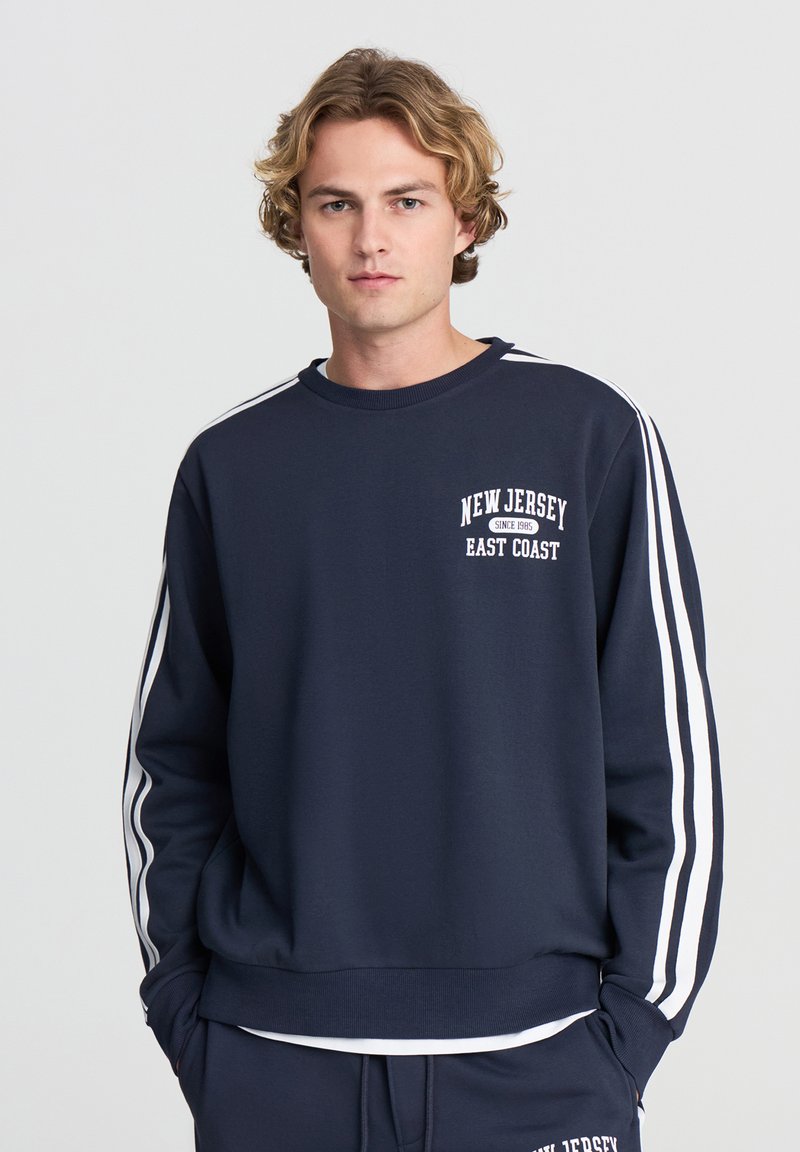 Felpa della marina in misto cotone con strisce bianche sulle maniche. Presenta una grafica "New Jersey East Coast" sul petto. Collo e polsini a coste.