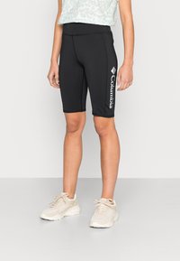 Černé elastické šortky s vysokým pasem, s bílým logem Columbia na levé straně. Boty jsou světle béžové s texturovanými povrchy.