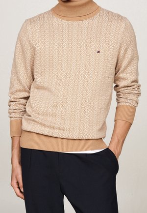 Pull à col roulé beige à motifs avec poignets et ourlet côtelés, porté avec un pantalon noir, main dans la poche.