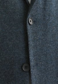 Tamno plavi č wool blejzer s teksturiranim herringbone uzorkom. Ima izrezani ovratnik i jedan crni gumb sprijeda.