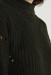 Pull vert foncé en tricot avec un col montant, un motif texturé et de petits trous dans le tissu, présentant un design épais.