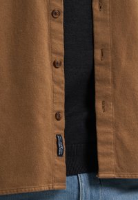 Bruine wollen button-up shirt met ronde bruine knopen, een labeltag en een gestructureerde afwerking, over een zwart kledingstuk gedragen.