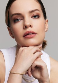 Złoty bransoletka o skręconym wzorze noszona na nadgarstku. Tło jest neutralne, podkreślając akcesorium i odcienie skóry.