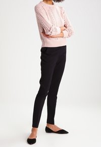 Pull en tricot rose clair avec un motif texturé, poignets côtelés, associé à un pantalon noir ajusté et des ballerines noires à bout pointu.