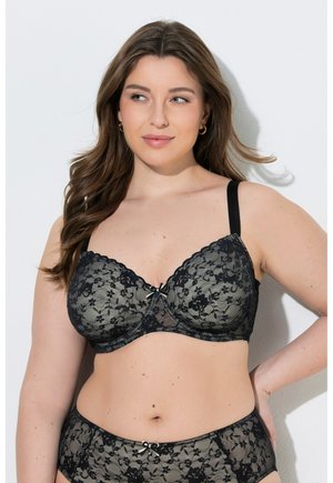 Woman with long brown hair wearing black lace bra and matching underwear, looking slightly to the side with hand on hip, against white background.