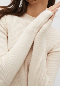 Haut beige clair à manches longues côtelé avec un col haut et des manches allongées. Texture douce, avec des rayures verticales sur toute la surface.