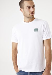 Camiseta de algodón blanca con mangas cortas, que presenta un logotipo rectangular verde en la zona del pecho izquierdo y un diseño de cuello redondo clásico.