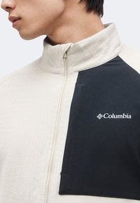 Άνδρας που φοράει μια μπεζ και μαύρη ζακέτα Columbia με φερμουάρ στον ψηλό γιακά, εμφανίζοντας το λογότυπο στο μαύρο μέρος στο στήθος.