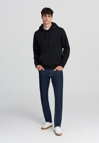Felpa nera in cotone con cappuccio regolabile, tasca frontale; abbinata a jeans in denim blu scuro e sneakers bianche. Design minimalista.