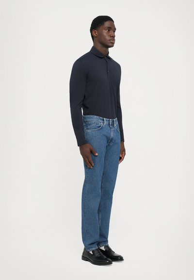 Man standing in dark long-sleeve polo, blue jeans, and black leather loafers against a plain light background.