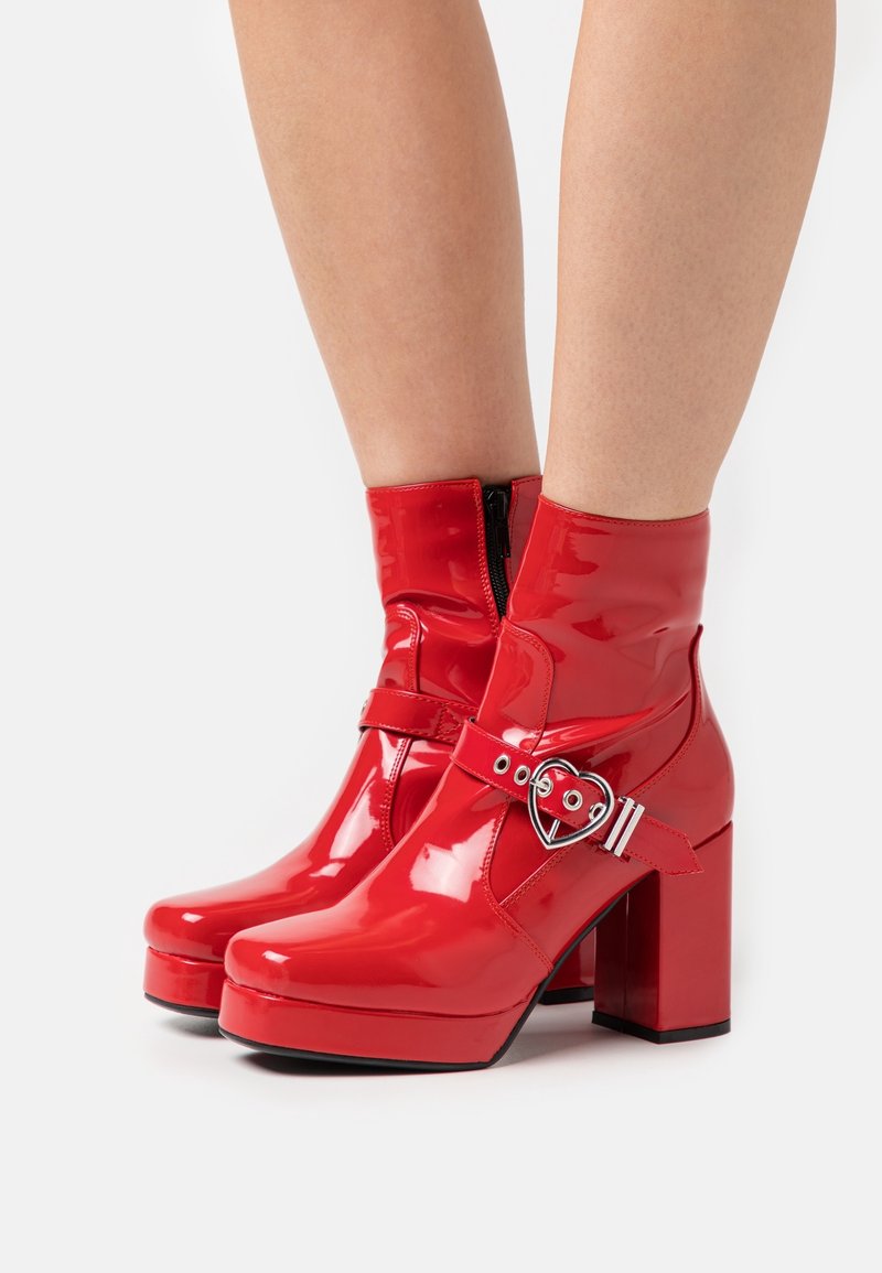 Bottines rouges en cuir verni brillant avec talons épais, semelles plateformes, boucles argentées en forme de cœur et fermetures éclair latérales, portées sur jambes nues.