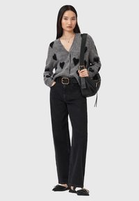 Cardigan grigio con motivi a cuore neri, con bottoni davanti, abbinato a jeans neri a vita alta e sandali flat neri, mentre tiene una borsa a spalla nera.