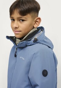 Blauwe waterdichte jas met capuchon, voorzien van een rits aan de voorkant, hoge kraag en een ronde zwarte logo patch op de mouw. Glad textuur.