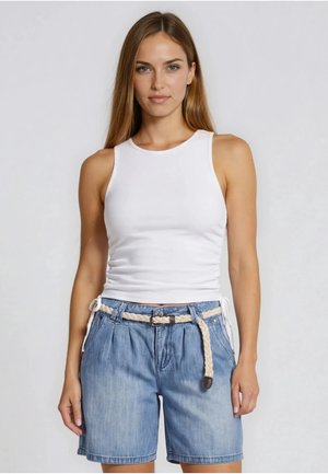 Kvinde iført en hvid ærmeløs top og lyseblå denimshorts med et flettet beige bælte, stående foran en ensfarvet baggrund.