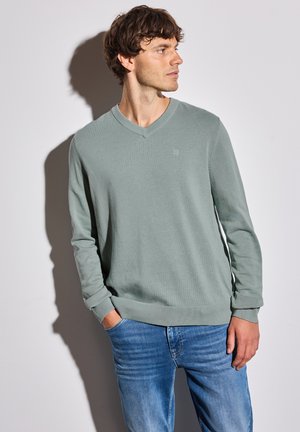 Light green, long-sleeve V-neck sweater made from soft fabric, featuring ribbed cuffs and hem, worn with blue jeans. Minimalist logo detail.