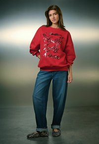 Bluza roșie cu inscripția "Holly Jolly Kinas" din paiete negre strălucitoare, purtată cu blugi lejeri albaștri și pantofi negri cu detalii decorative.