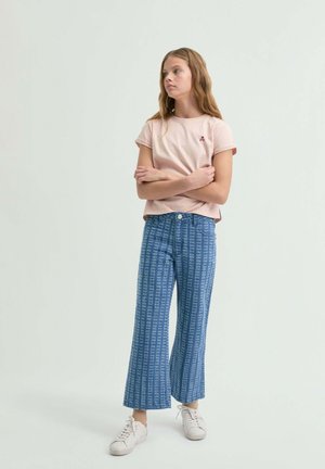 Joven de pie con los brazos cruzados, vistiendo una camiseta rosa claro, jeans recortados azules con patrón y zapatillas blancas contra un fondo liso.