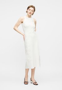 Femme portant une robe midi blanche sans manches avec des détails en dentelle florale, posant avec les mains derrière le dos, et des sandales à talons noires à lanières.