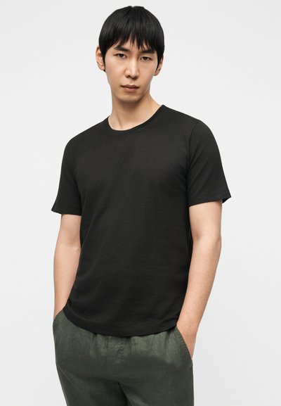 Μαύρο κοντομάνικο μπλουζάκι με στρογγυλή λαιμόκοψη. Λείος υφής και άνετη εφαρμογή. Το μοντέλο φοράει πράσινο λινό παντελόνι. Απλός σχεδιασμός χωρίς ορατά λογότυπα.