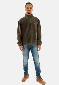Sudadera de forro polar verde olivo con cuello de media cremallera y detalle de botón. Presenta un pequeño logotipo en el pecho, combinada con jeans de color azul claro y zapatos marrón claro.